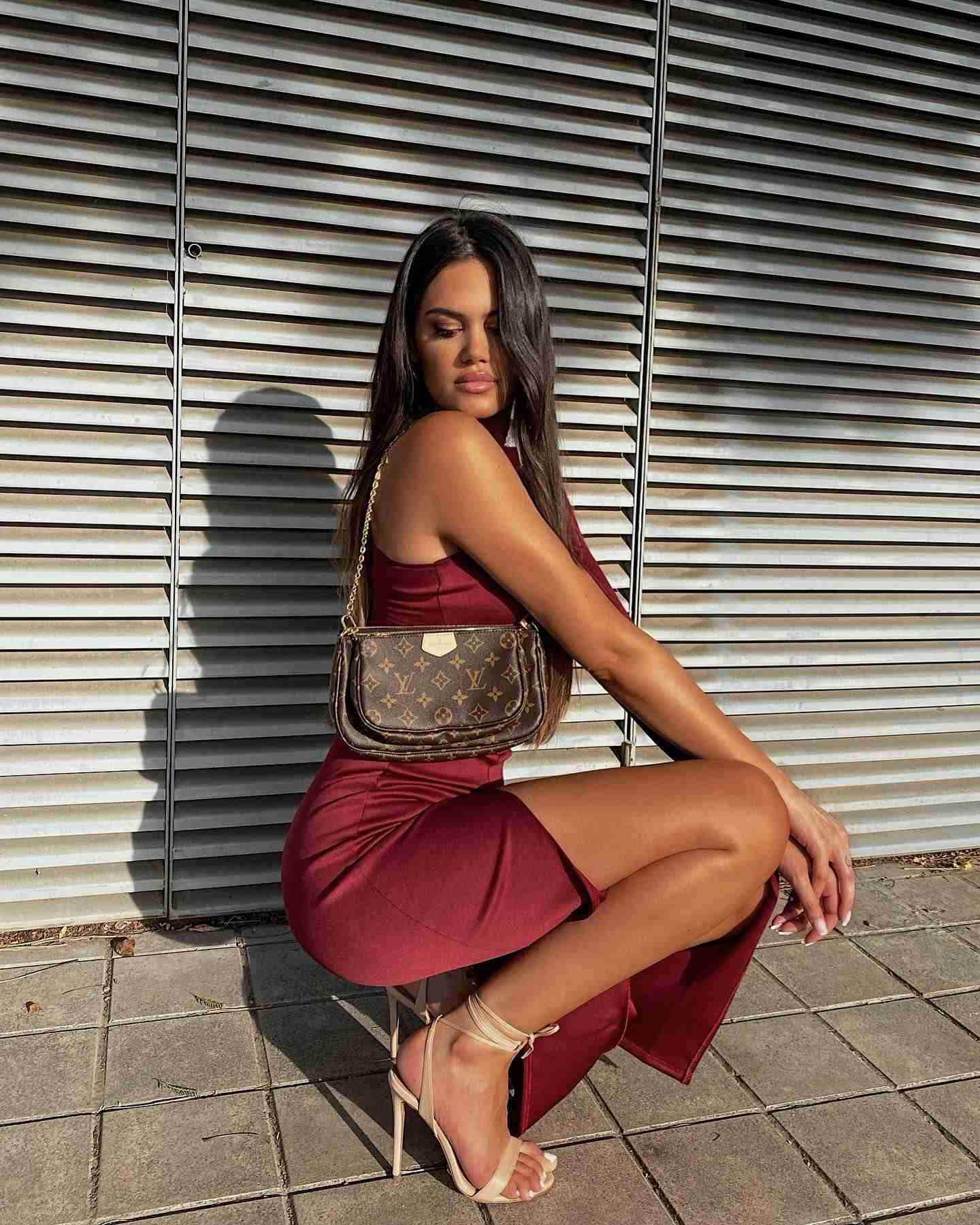 Mujer en posicion sentada llevando un vestido largo color burdeos con abertura lateral, un bolso y unas sandalias de color beige